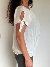 Afbeelding in Gallery-weergave laden, Witte Top Dobby heeft een kort mouwtje met een strikje, wat een leuk detail is. De top heeft een ronde hals, een rij knoopjes en heeft aan de onderzijde een elastische band met ruffles. Top kan zowel op de schouder als off-shoulder gedragen worden en is verkrijgbaar in verschillende kleuren. Kleur : Blauw, Geel, Wit