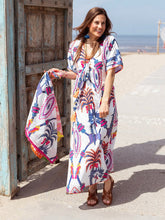 Afbeelding in Gallery-weergave laden, Maxi Dress Rosa Blue Bird Long van Place du Soleil in de kleurstelling wit, blauw en fuchsia. De Rosa Blue Bird Long Dress is een prachtig uitlopende lange gevoerde jurk met steekzakken en heeft een smallere taille met (vaste) ceintuur met oranje fluo flosjes. Jurk heeft een V-hals met fluo oranje borduursel en korte mouwen, is afgezet met subtiele pailletten en is bewerkt met prachtig lurex draad.
