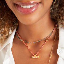 Load image into Gallery viewer, Kleurrijke ketting van rood en roze touw en gouden details. De gouden details zoals de bedel zijn gemaakt van roestvrij staal.
