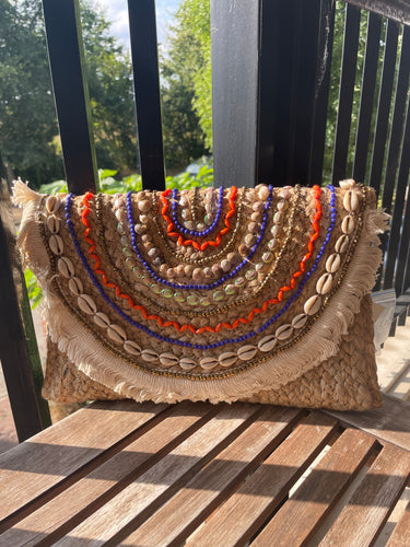 Clutch van Sisal, Boho Stijl, bewerkt met schelpen, gekleurde kraaltjes en korte franjes. De clutch is gevoerd, heeft een lang hengsel en is sluitbaar met klep en magneetsluiting. Prachtige applicatie! Deze clutch is verkrijgbaar in verschillende applicaties: Roze en Oranje/paars.